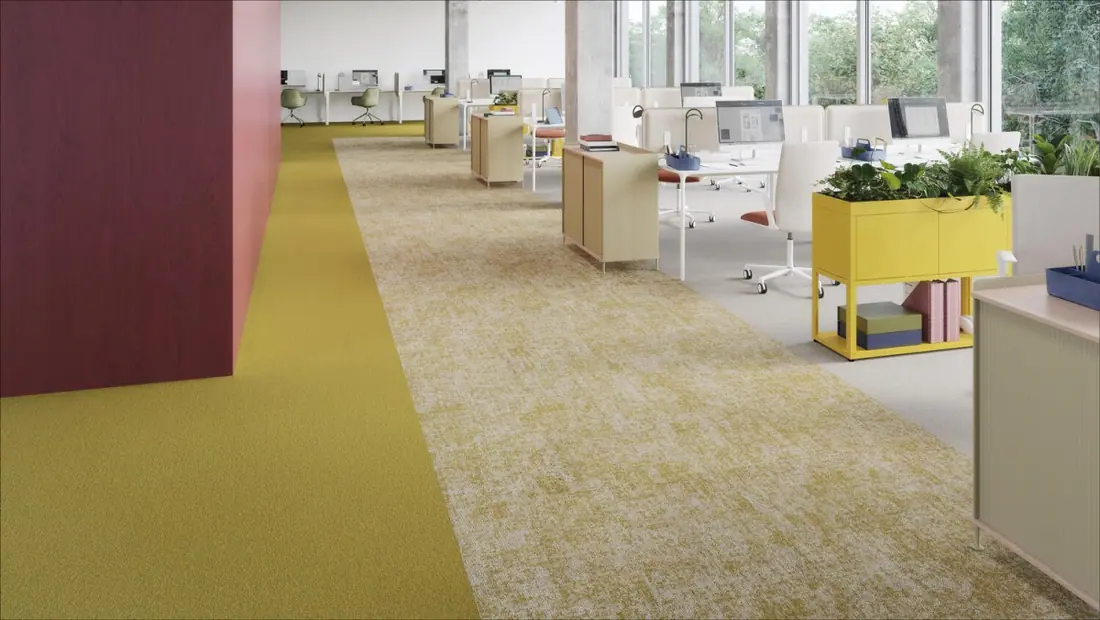 Open kantoorruimte met Desso Essence Roots en de Desso Essence Pure tapijttegels. De vloer is verdeeld in twee secties: een rijke, mosterdgele tapijttegels links en een mosterdgele Desso Essence Roots met subtiele patronen in het midden. Moderne bureaus met computers en lage houten scheidingswanden vullen de ruimte. Planten zorgen voor een vleugje groen, terwijl grote ramen rechts natuurlijk licht binnenlaten, wat de textuur en kleuren van de Desso tapijttegels benadrukt.