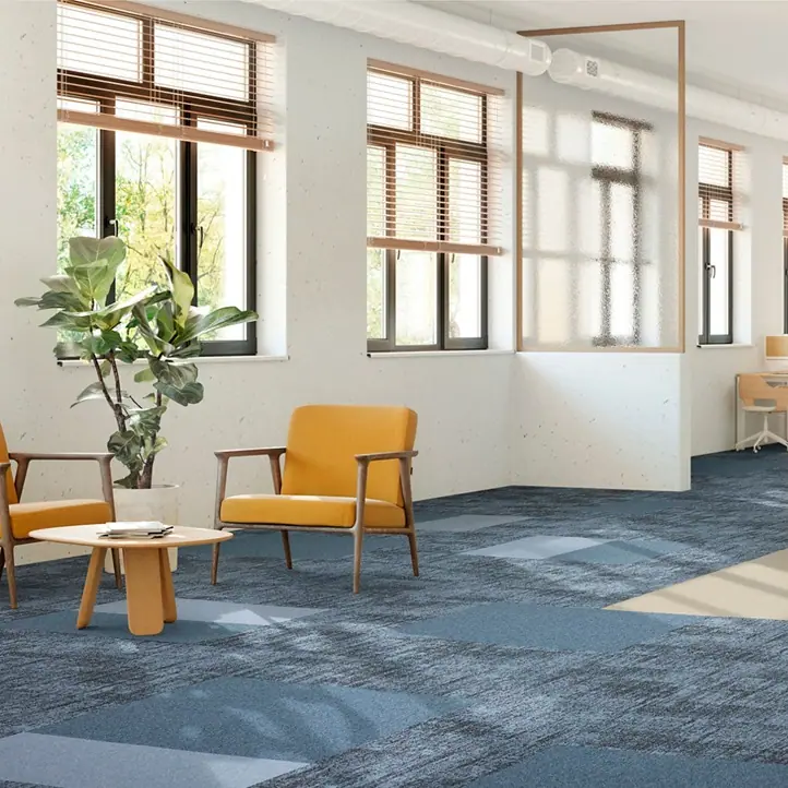 Modern kantoorinterieur met Mohawk Reflect 559 tapijttegels in verschillende tinten blauw en grijs. De ruimte bevat comfortabele geel oranje fauteuils met houten armleuningen, gegroepeerd rond een ronde lage houten tafel. Grote ramen met houten jaloezieën laten veel natuurlijk licht binnen, wat reflecteert op de lichte muren. Een grote groene plant in een decoratieve pot voegt een natuurlijk element toe. De combinatie van kleuren, materialen en licht creëert een frisse, uitnodigende werksfeer die zowel rustgevend als stimulerend is.